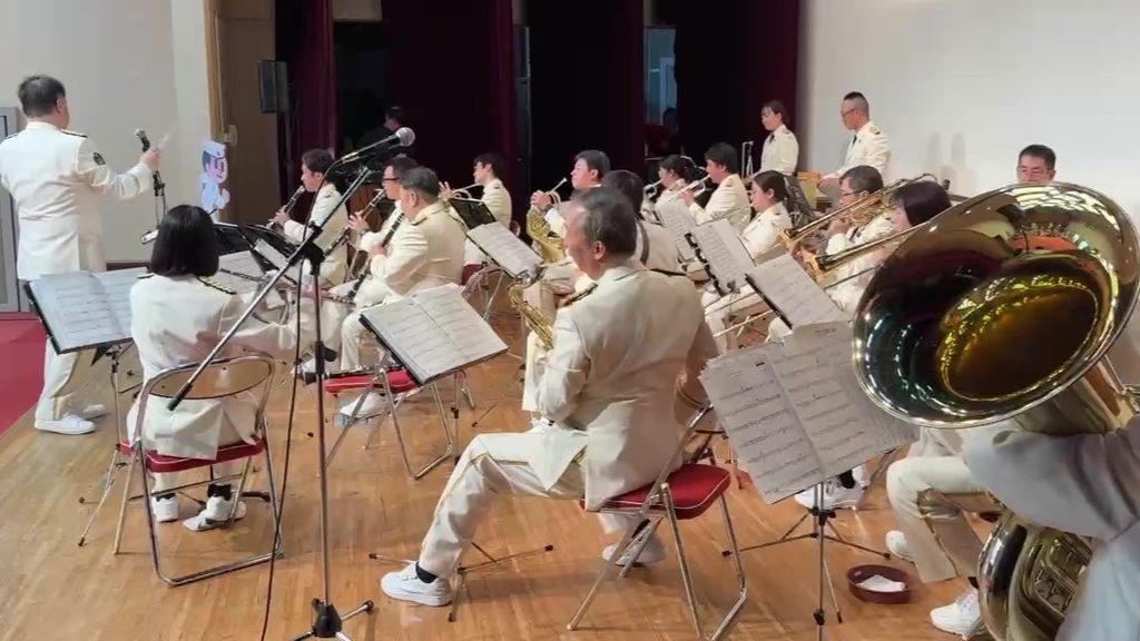 屋内ステージで白い制服を着た吹奏楽団(滋賀県警察音楽隊)が演奏し、ステージ手前に収音用マイクが設置されている様子。滋賀県東近江市のやわらぎホールで開催されたイベントにて。