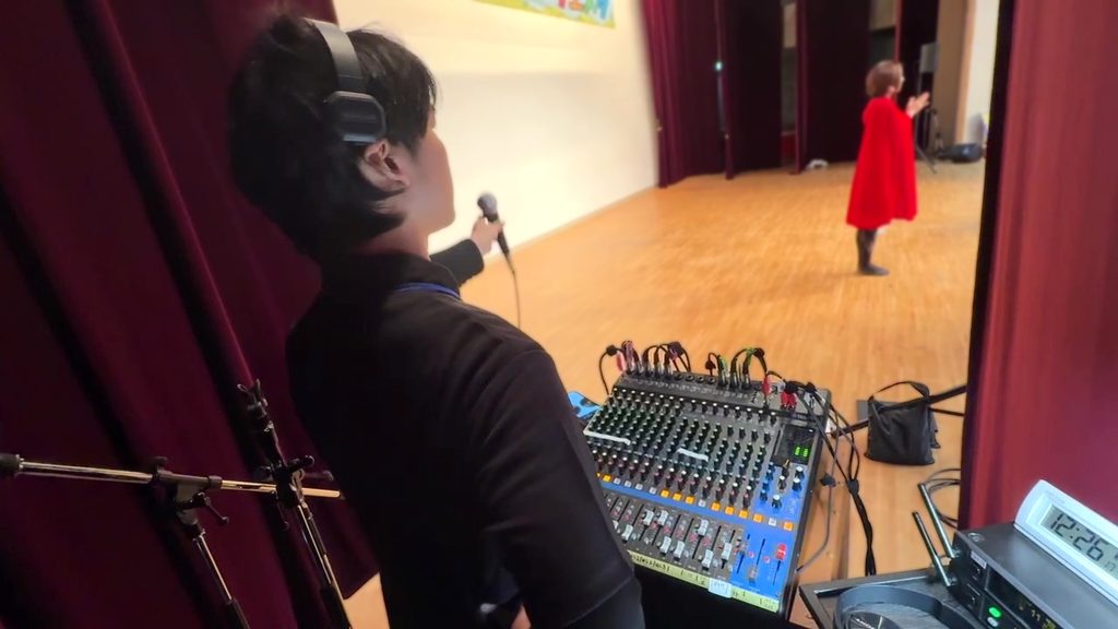 舞台袖のミキサー卓横で、ヘッドホンを装着したスタッフがマイクを持ち、ステージ上の出演者と連携をとっている様子。滋賀県東近江市のコミュニティセンターにて開催された能登川ふれあいフェスタにて。