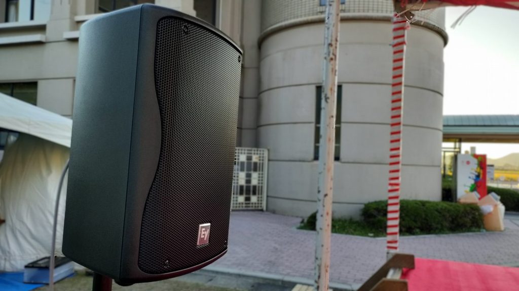 滋賀県近江八幡市のフェスタ現場屋外ステージ脇に設置された、Electro-Voice（エレクトロボイス）製のコンパクトなモニタースピーカー「ZX1」。背景には会場の建物やテント、紅白幕が設置されたステージが写っている。