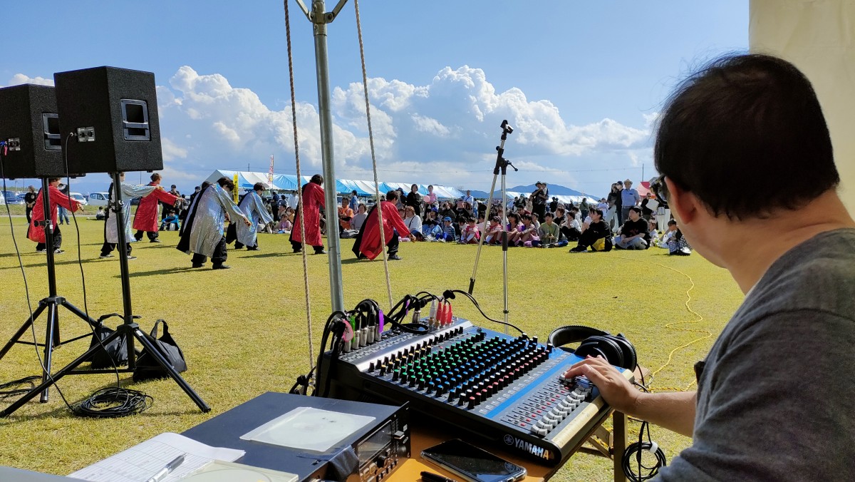晴天の野外フェスタ会場にて、ミキサーを操作しながら芝生ステージの演目を音響オペレートする様子。滋賀県大津市の仰木西公園にて開催された仰木の里フェスタにて。