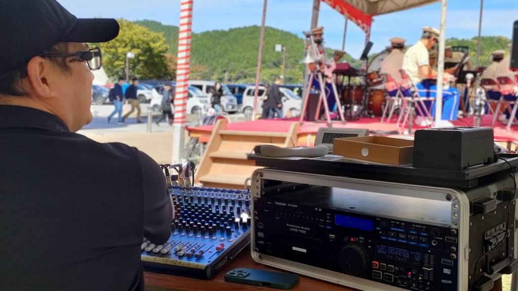 滋賀県近江八幡市で開催された屋外フェスタの音響オペレート席。黒いキャップとポロシャツを着用したスタジオ トイボックスのPAオペレーターが、左手にあるYAMAHAミキサーや右手のラック機材（TASCAM CDプレーヤー等）を前に、ステージで行われている演奏や進行を真剣に見つめている後ろ姿。背景には赤い絨毯のステージと演奏者、屋外に集まる来場客や車、山々が写っている。